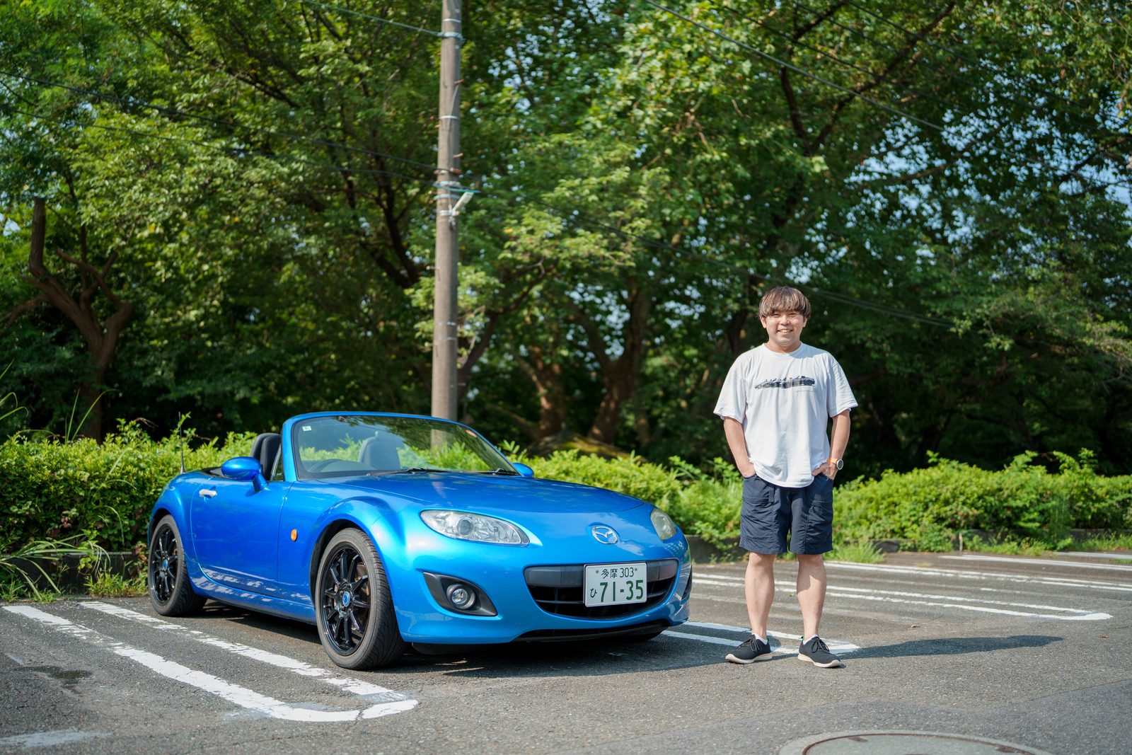 青いマツダ・ロードスター（NC）の横に立つ木下さん。街路樹を背景にした駐車場での全身ショット